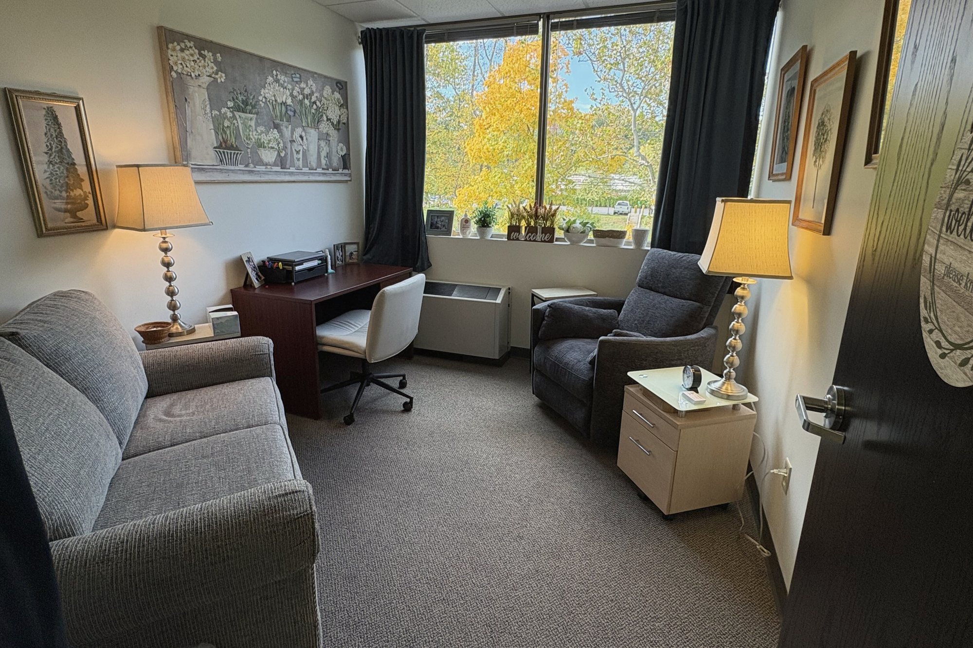 A therapy room at the Shelton office with a gray sofa, an armchair and a window onto autumn trees